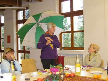 Ökumenisches Frauenfrühstück Frauen sitzen an einem schön gedeckten Frühstückstisch. Eine Frau mit einem geöffneten Schirm in der Hand erzählt etwas, das die anderen zum Lachen bringt